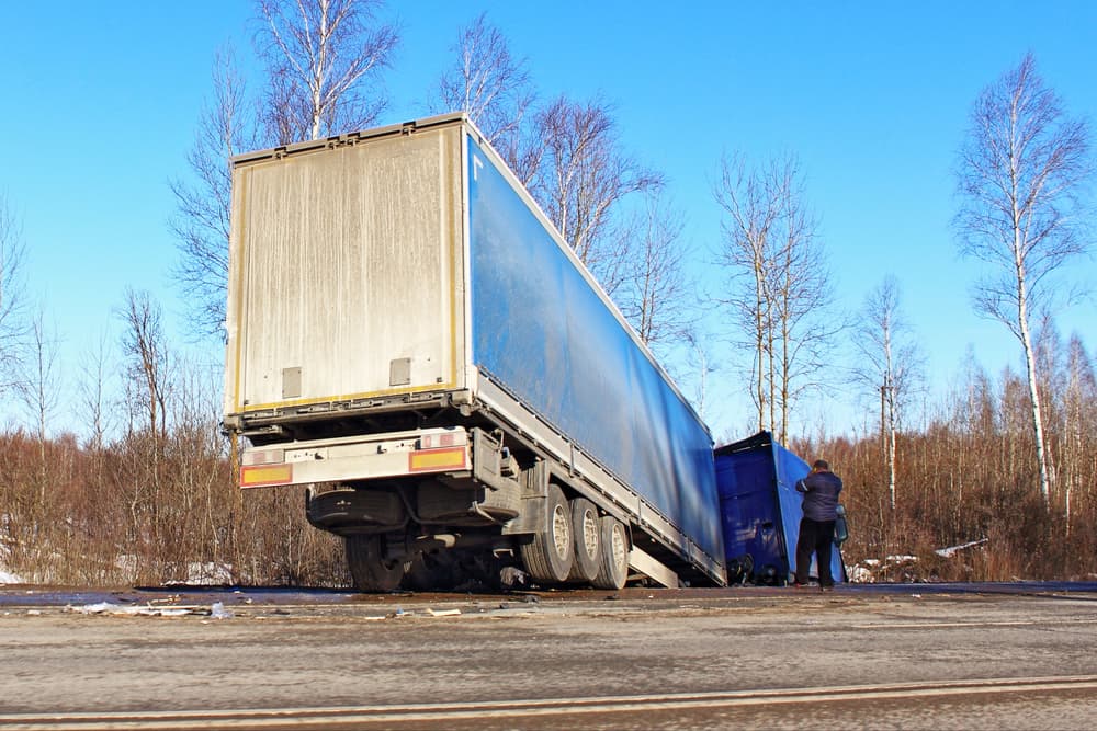 A semi-truck lies tilted and wrecked in a roadside ditch on a sunny spring day, the scene of an accident on a suburban highway.