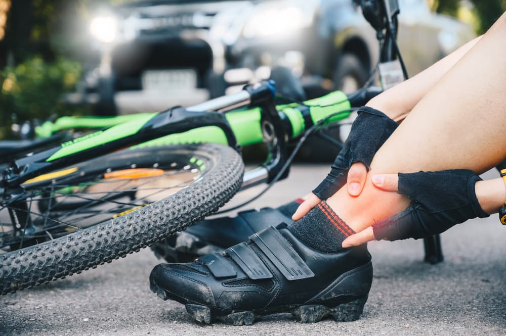 Cropped shot of cyclist woman suffering from ankle pain after car accident on the road.